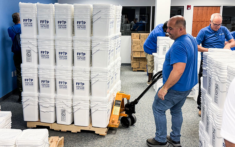 A group of men in blue shirts stands around a pallet stacked with white containers. 